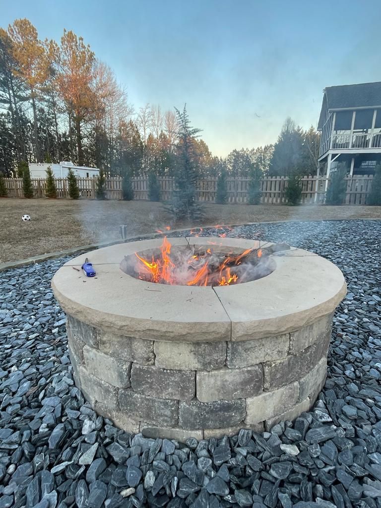 Fire burning in a stone fire pit outdoors, surrounded by dark gray stones, with a house and trees in the background.