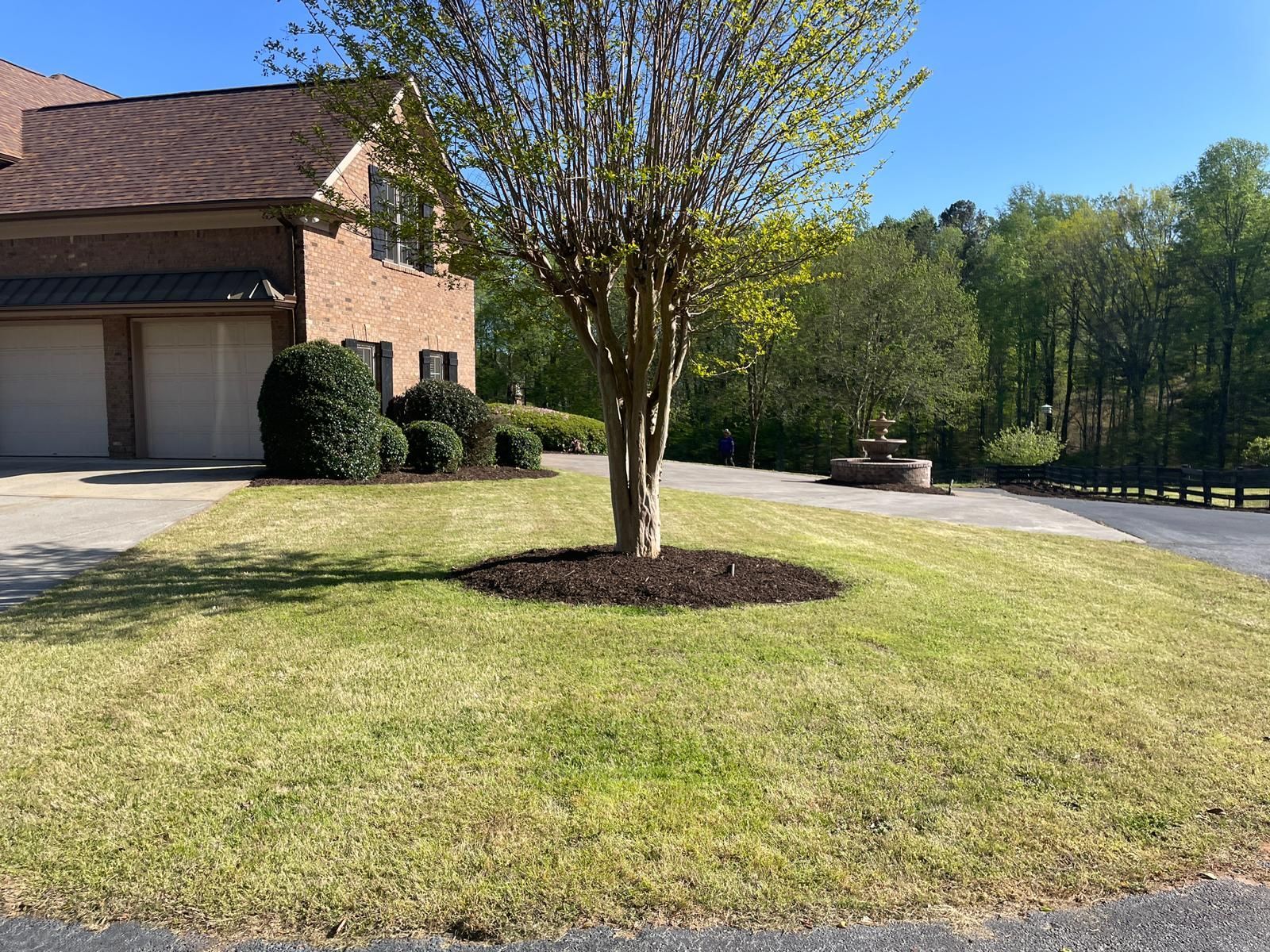 Green lawn in front of a brick house with a tree circled with mulch and a stone fountain.