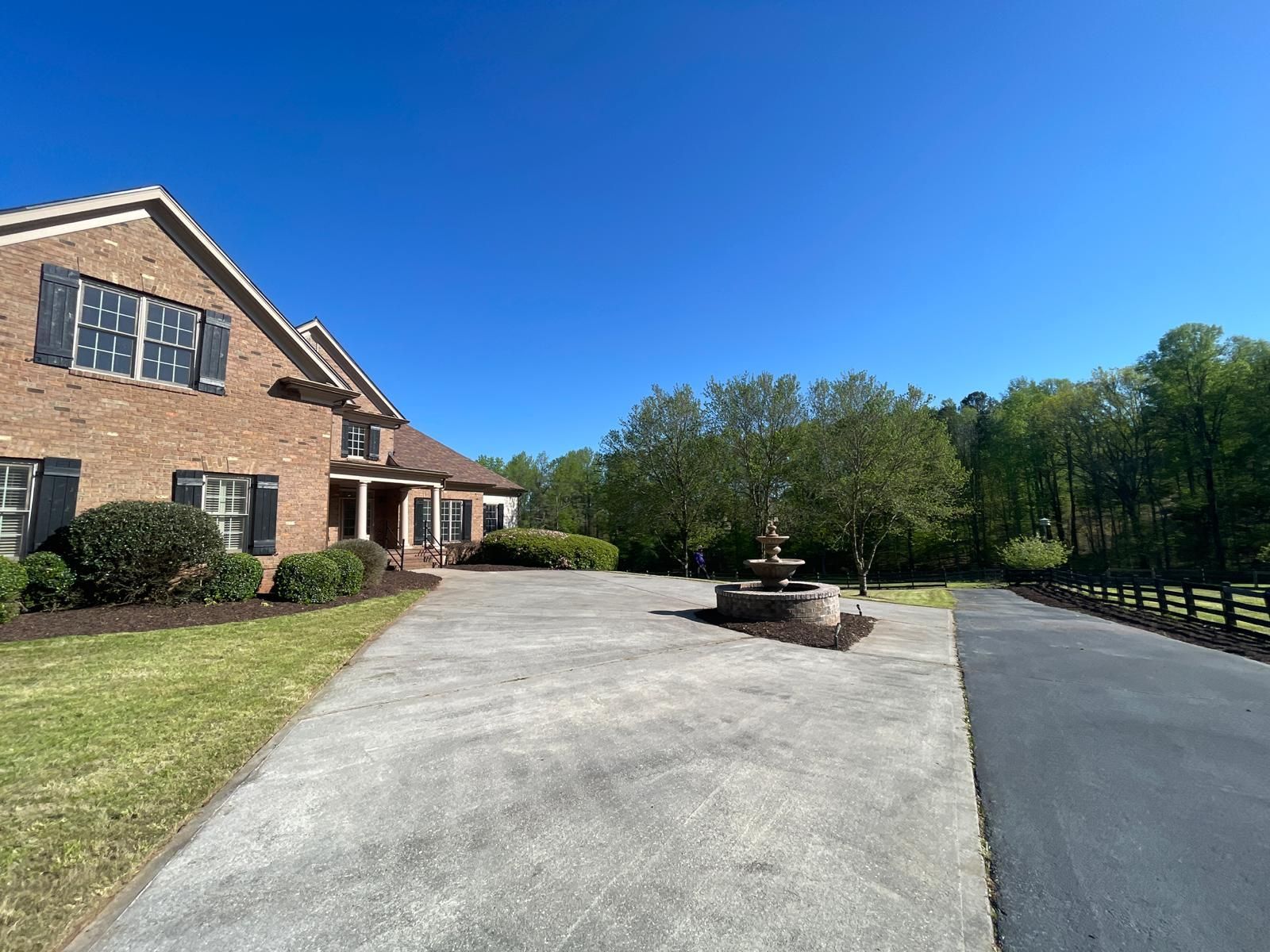 Brick house with a long driveway and fountain, under a clear blue sky.