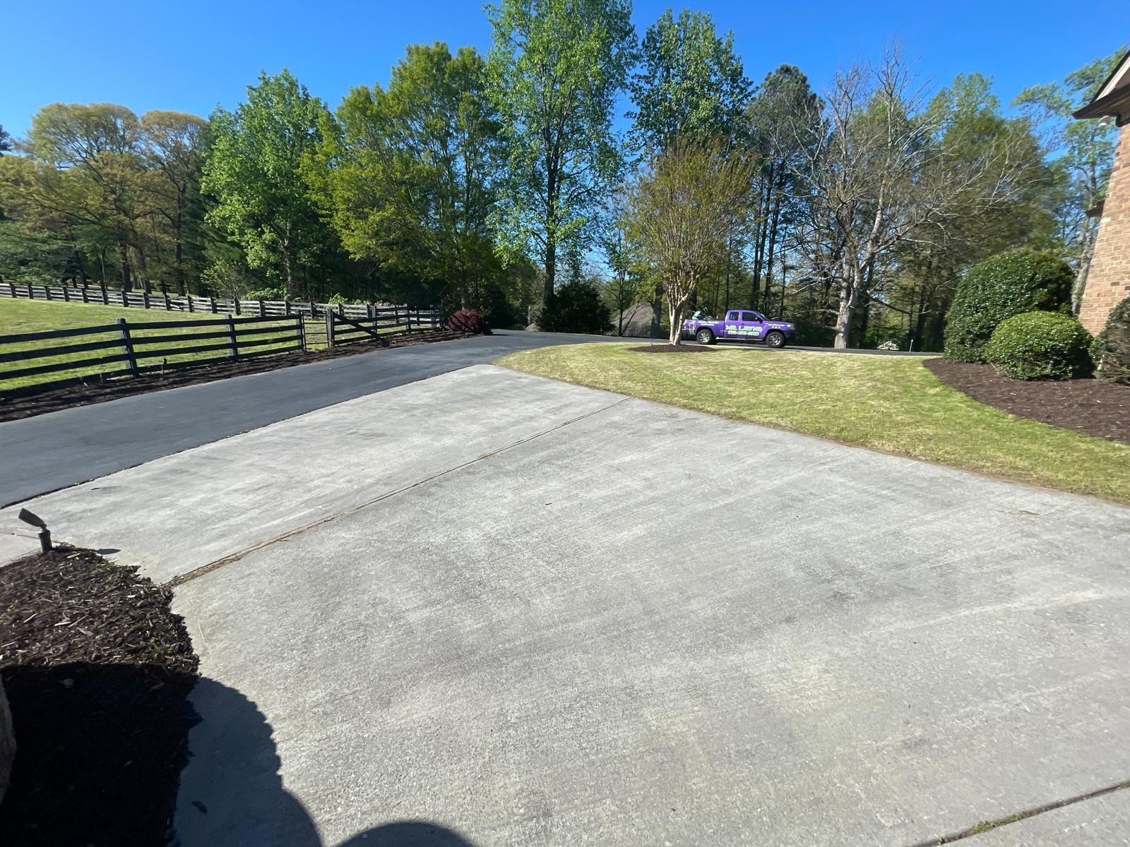 Concrete driveway leading to grassy area with car, trees, and wooden fence. Bright sunny day.