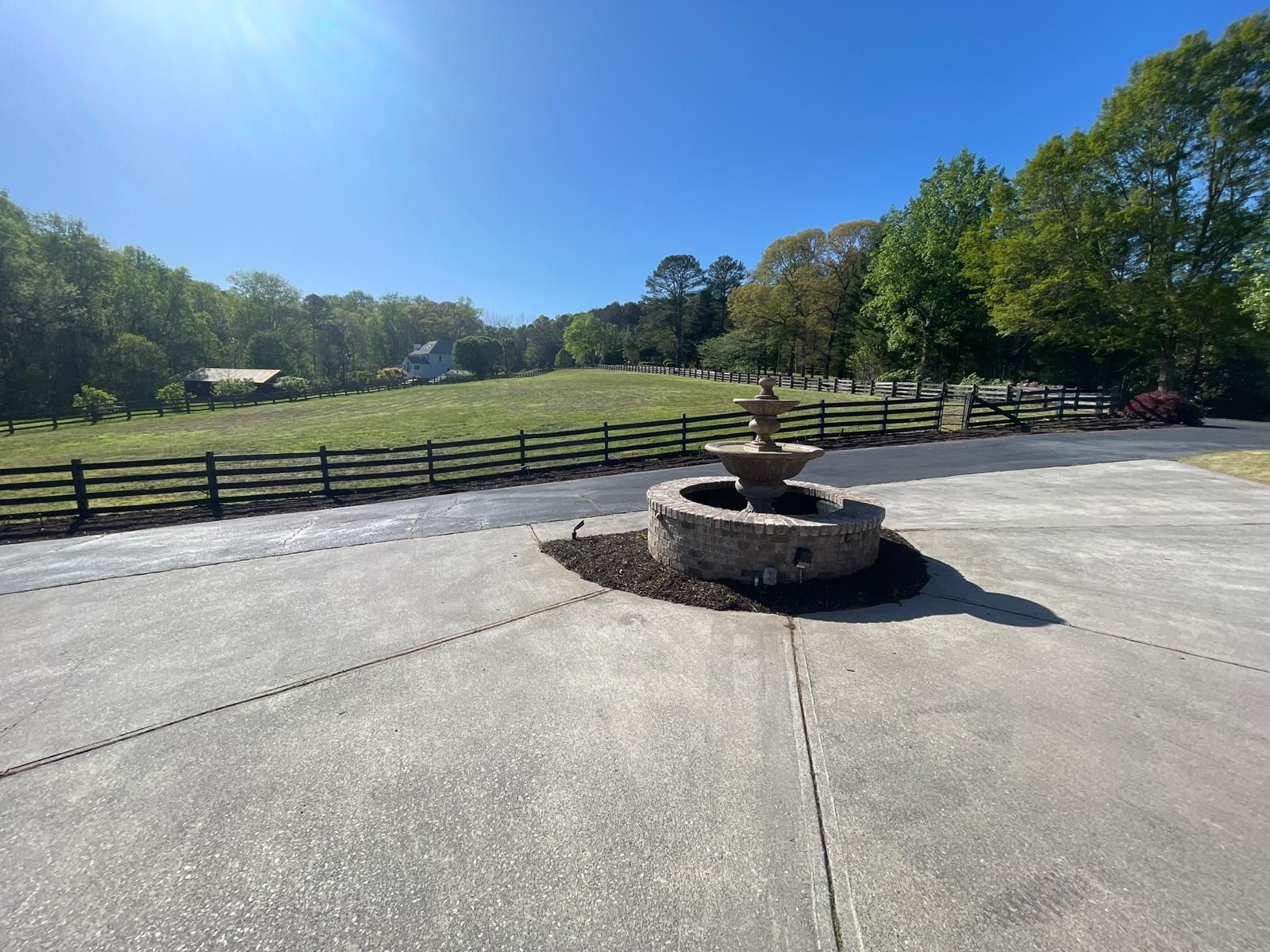 Stone fountain on a concrete patio, with a green field and trees in the background under a blue sky.