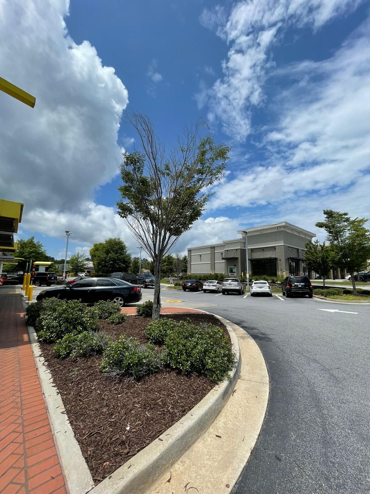 Drive-thru of a McDonald's restaurant with a tree and parked cars under a partly cloudy sky.