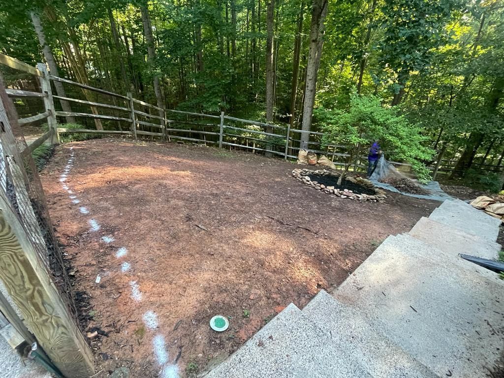 A backyard with a fence, mulch ground, stairs, and trees. Someone is gardening.