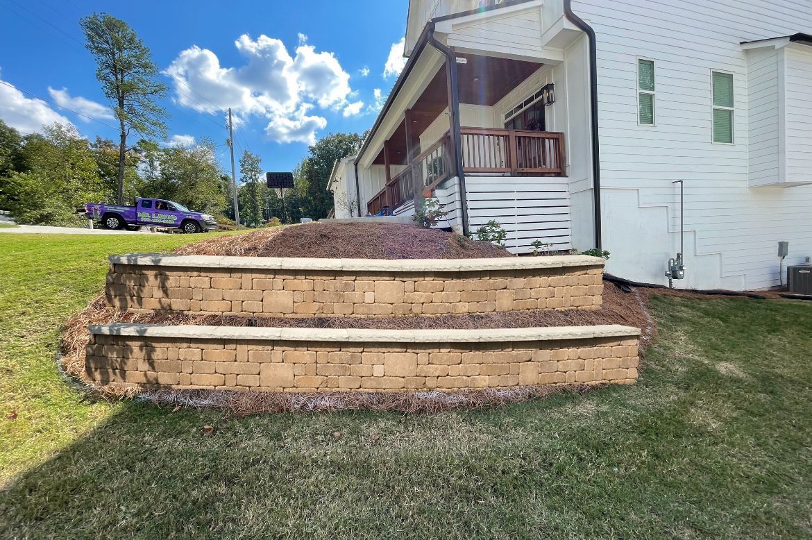 Retaining wall steps in front of a white house with a porch, surrounded by grass and mulch. Purple truck visible.