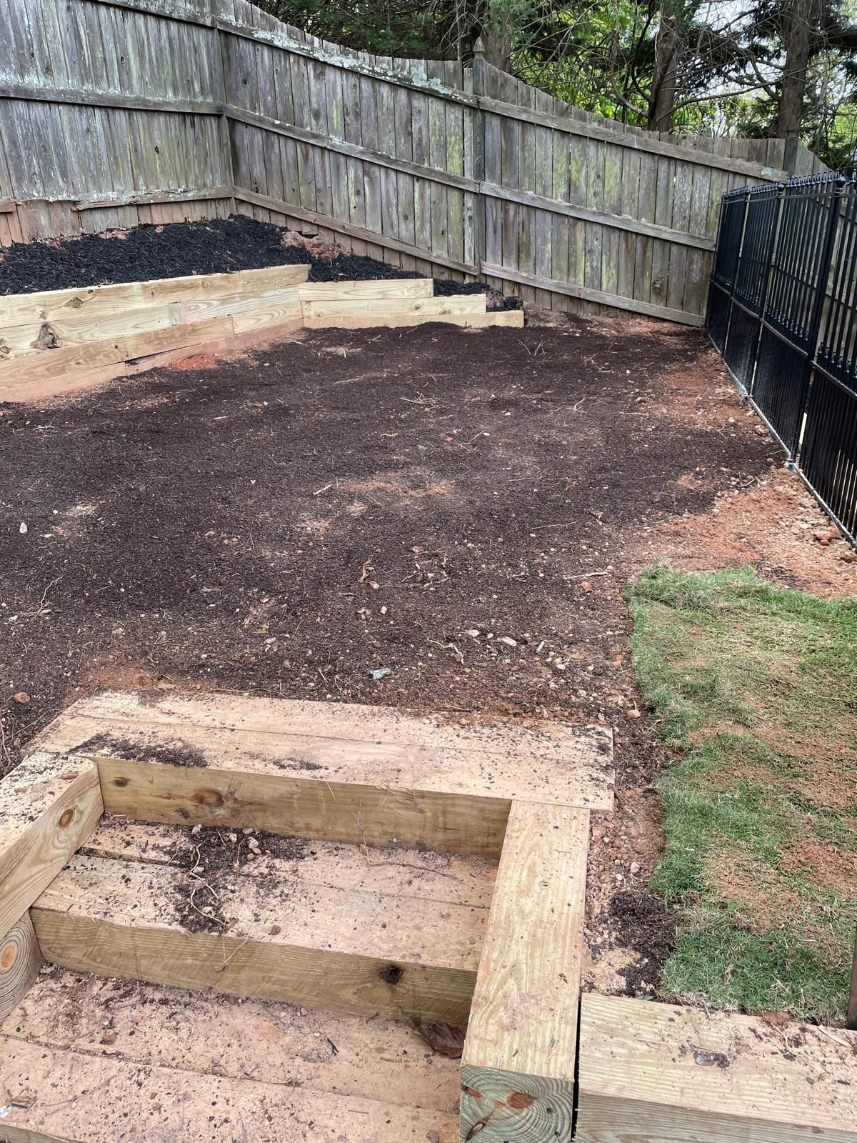 A backyard with steps, retaining walls, mulch, and fencing. Green grass is visible.