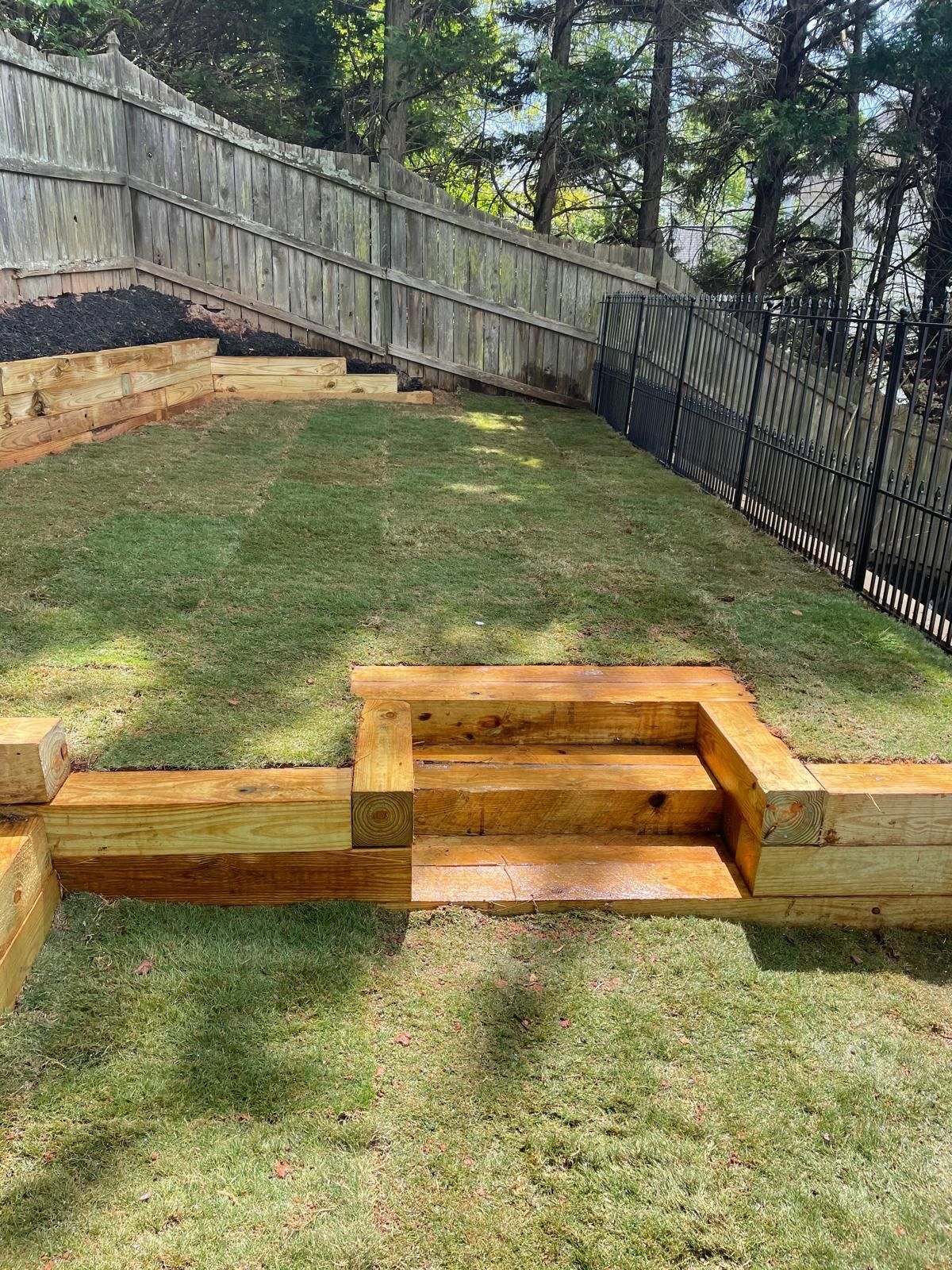 Lawn with wooden steps and retaining walls on a sloped yard. A fence and trees are visible in the background.