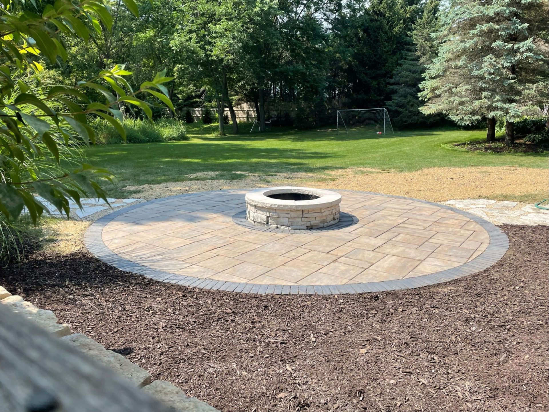 Circular stone patio with fire pit, surrounded by mulch, overlooking a grassy yard.