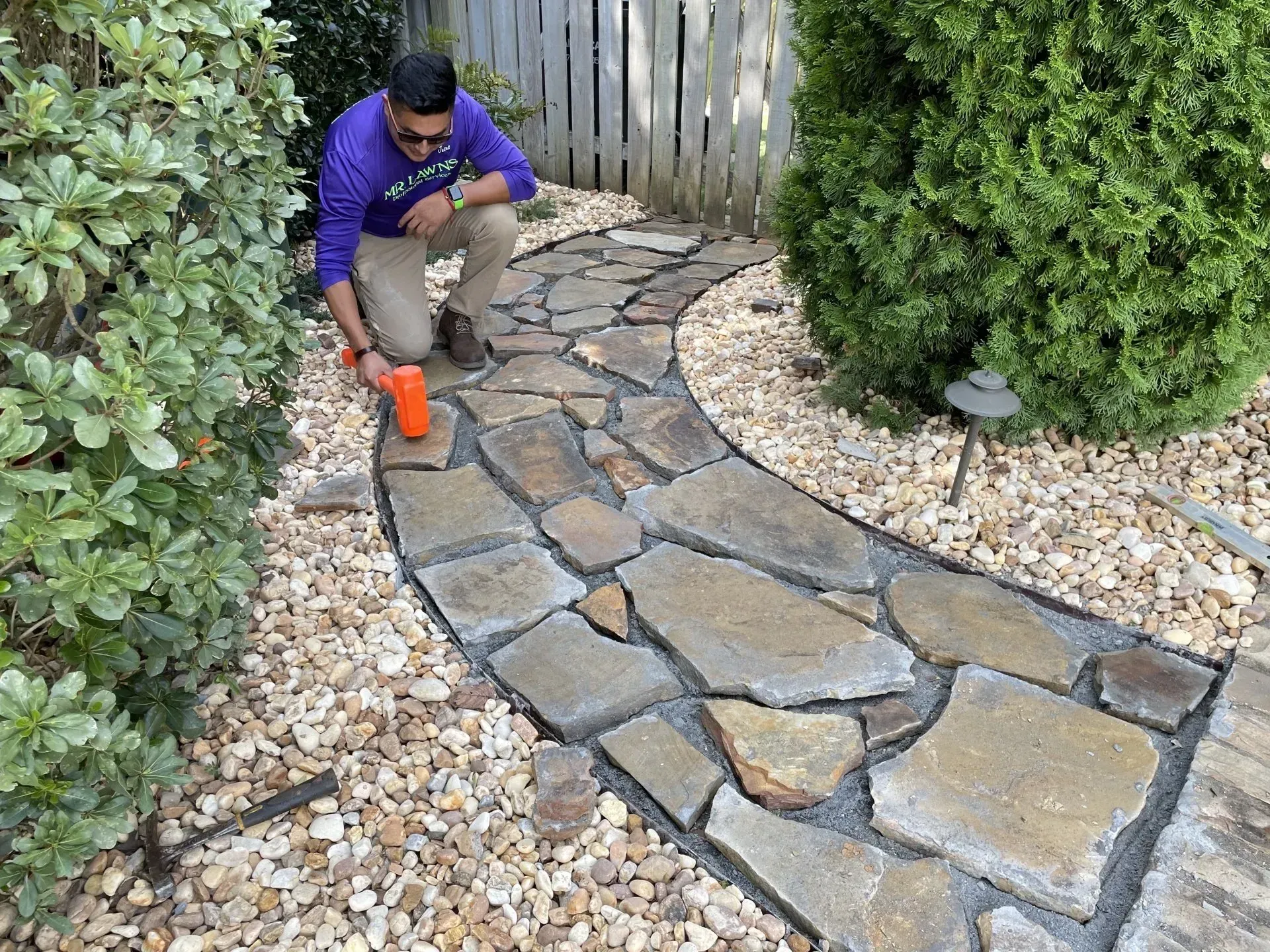 Person kneeling, placing stone in a pathway using a mallet. Pathway curves through yard with foliage and fence.