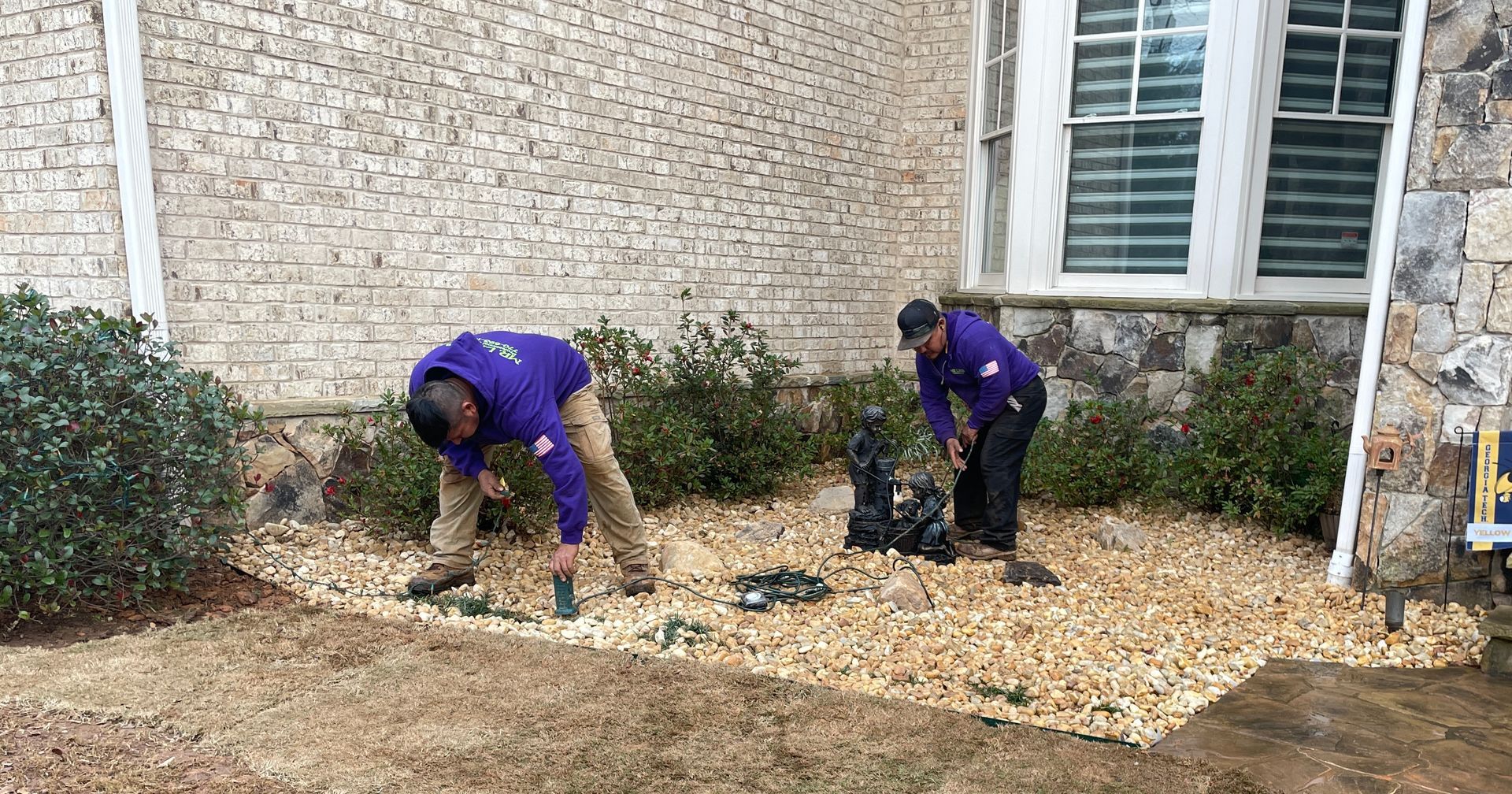 Two people in purple shirts work on a rock bed near a building.
