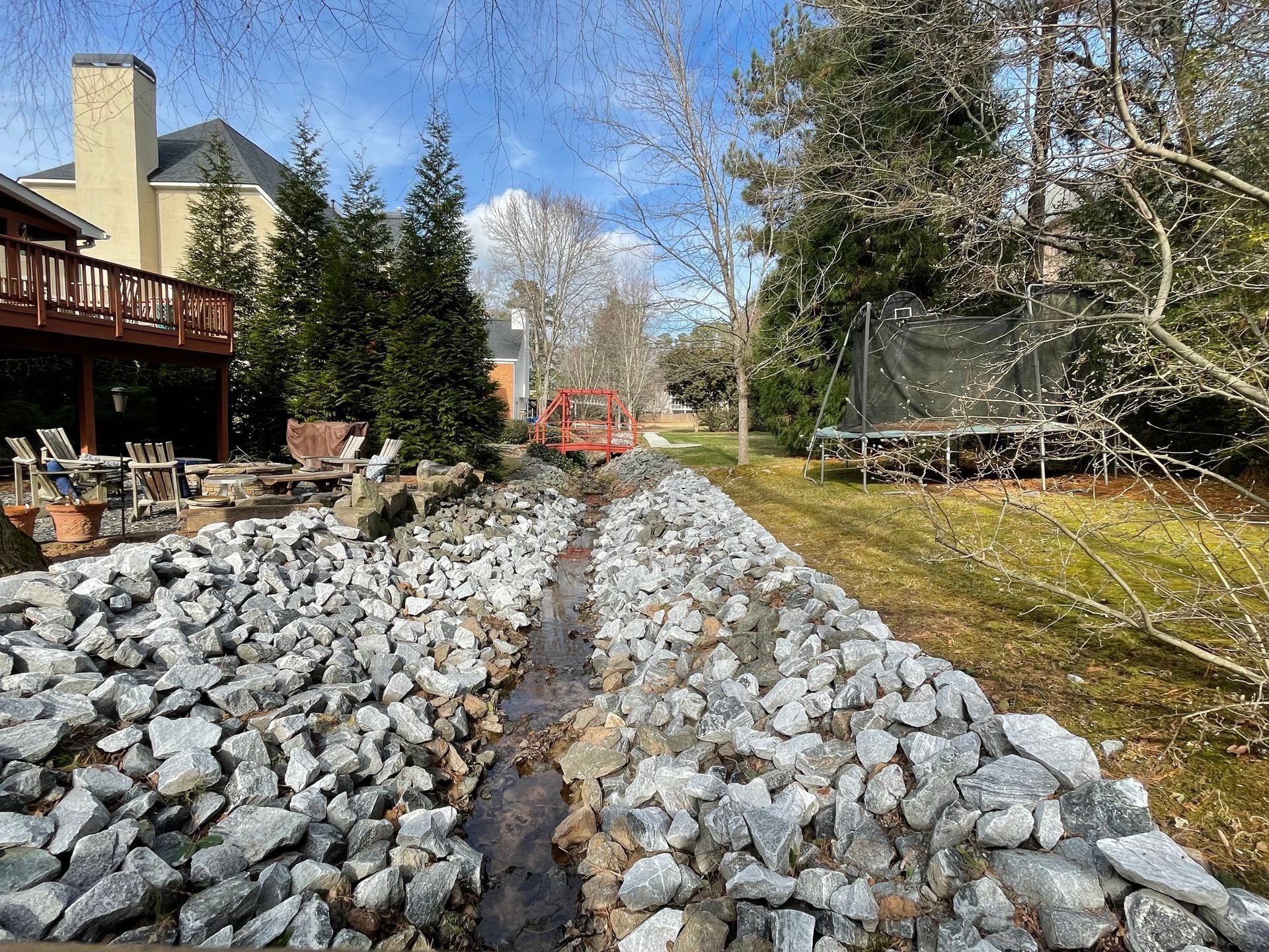 Rock-lined drainage ditch in a backyard, with a deck on the left and a trampoline on the right.