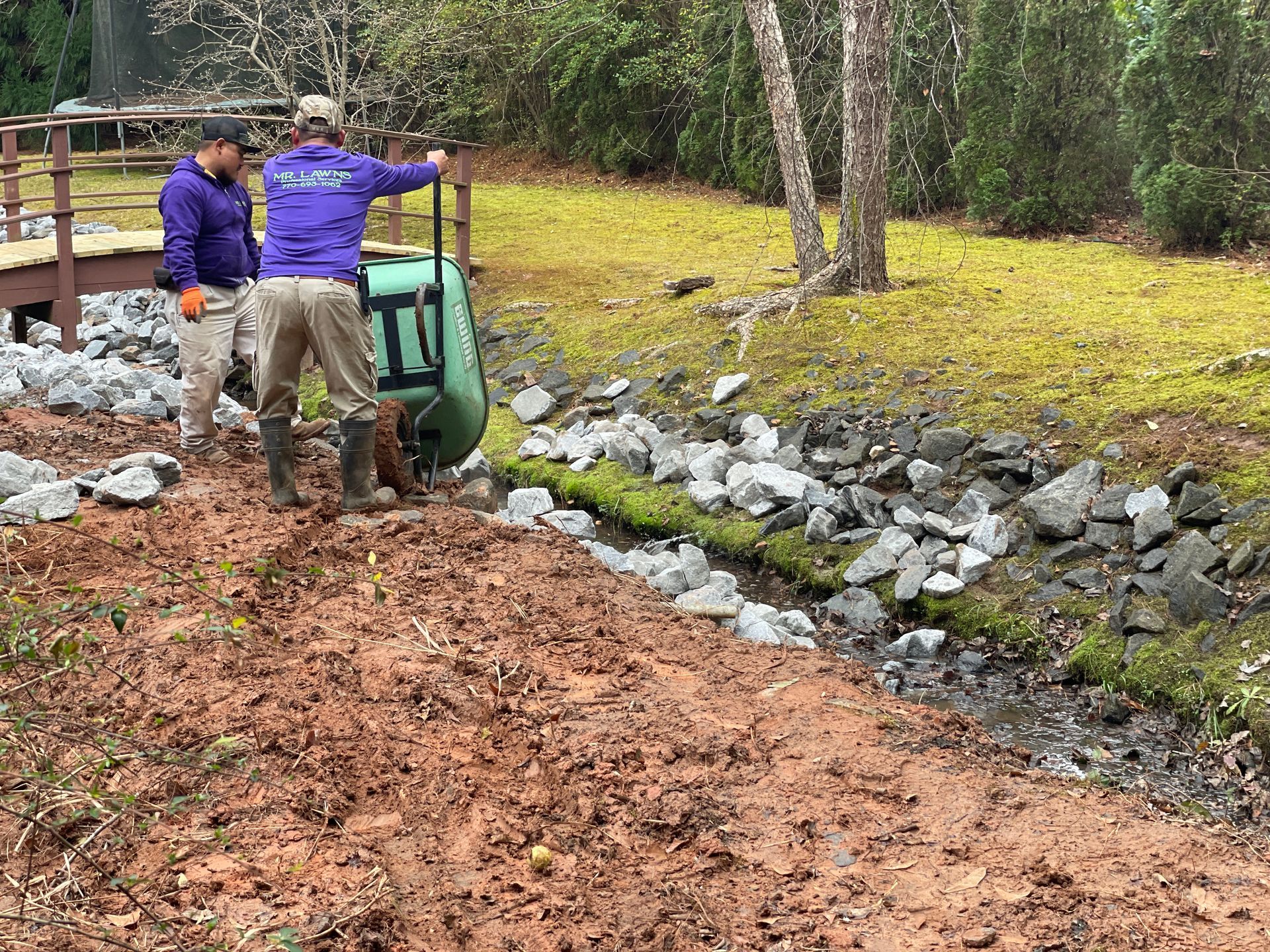 Two workers near a rocky stream bed, one with a wheelbarrow, working outdoors.