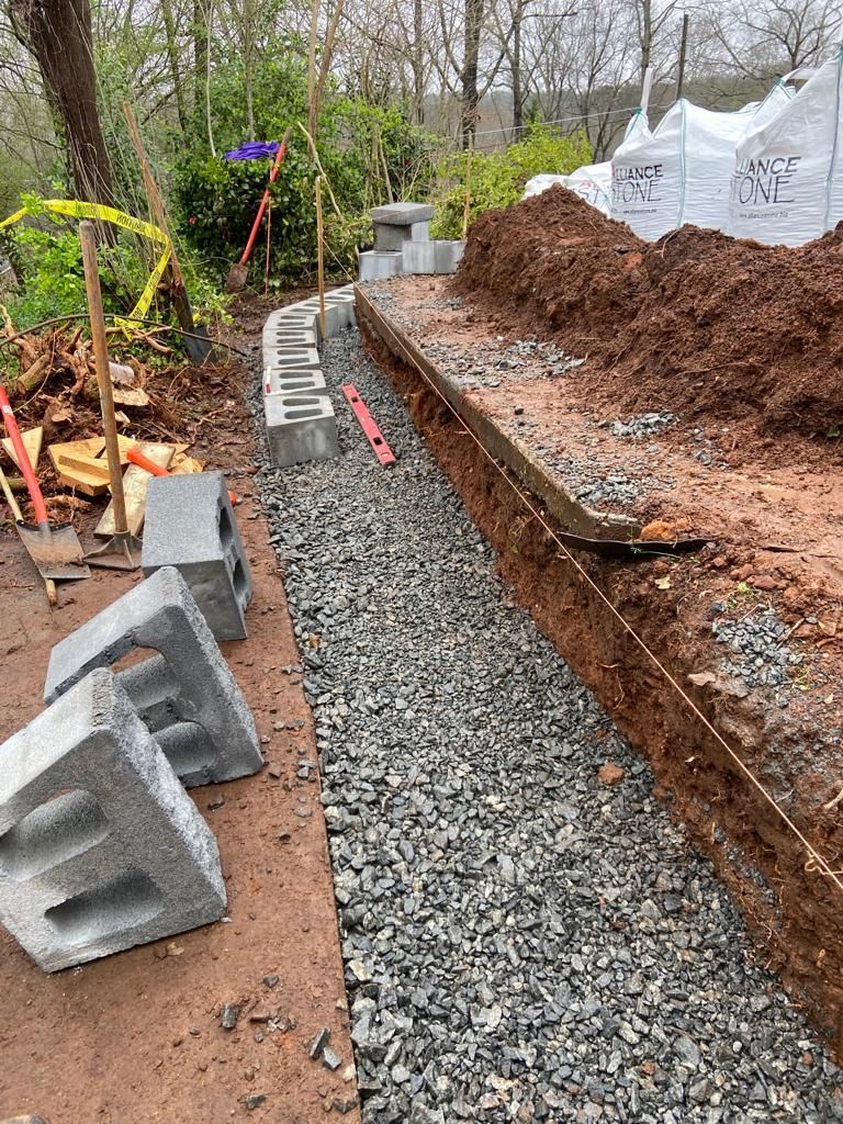 Construction site. Gravel-lined trench with retaining wall blocks. Soil pile, wooden beam, and leveling tools are present.