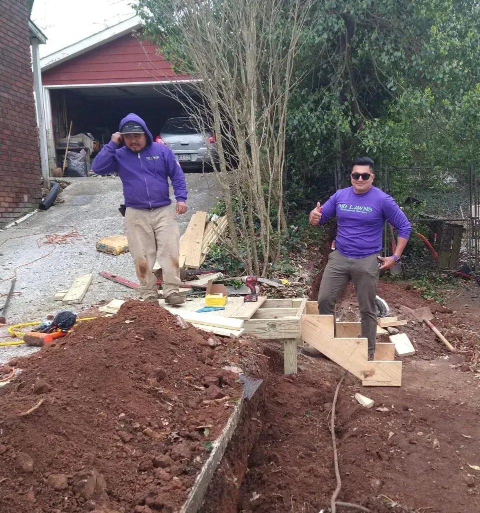 Two men in purple shirts work on a landscaping project. One gives a thumbs up. Dirt and tools are visible.