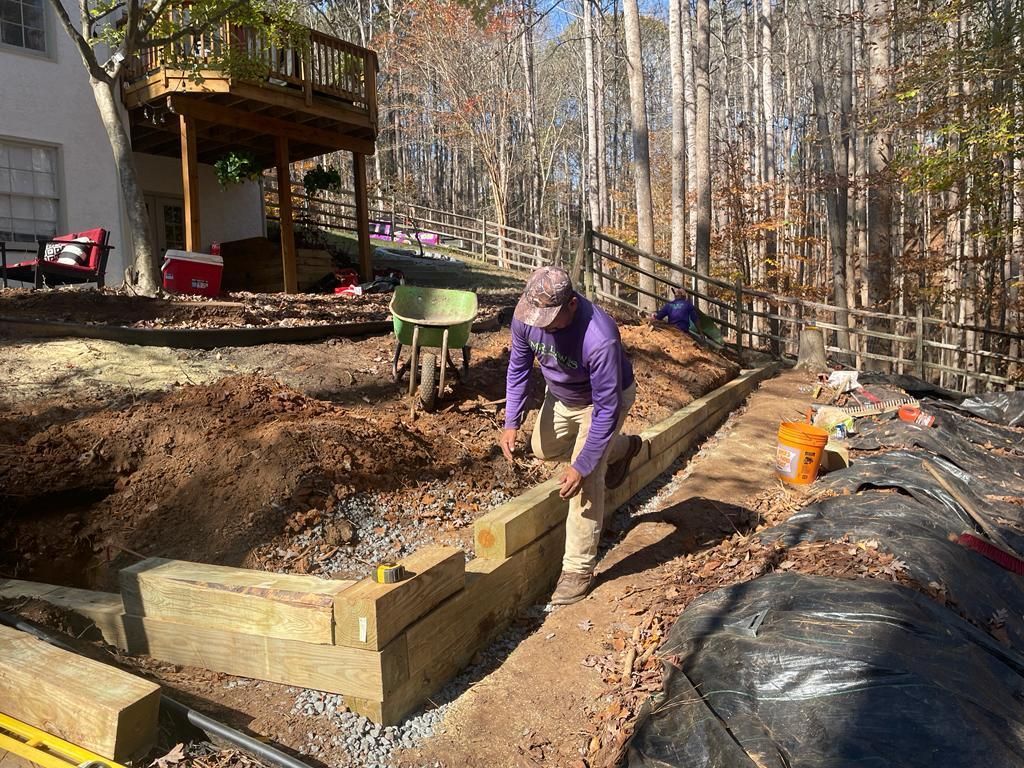 Man building a retaining wall with wooden beams on a hillside with trees and house in the background.