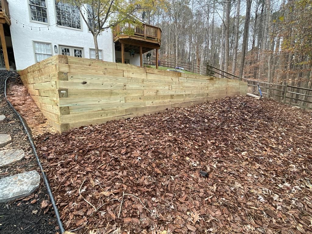 Wooden retaining wall next to a house with a deck, covered in mulch and leaves.