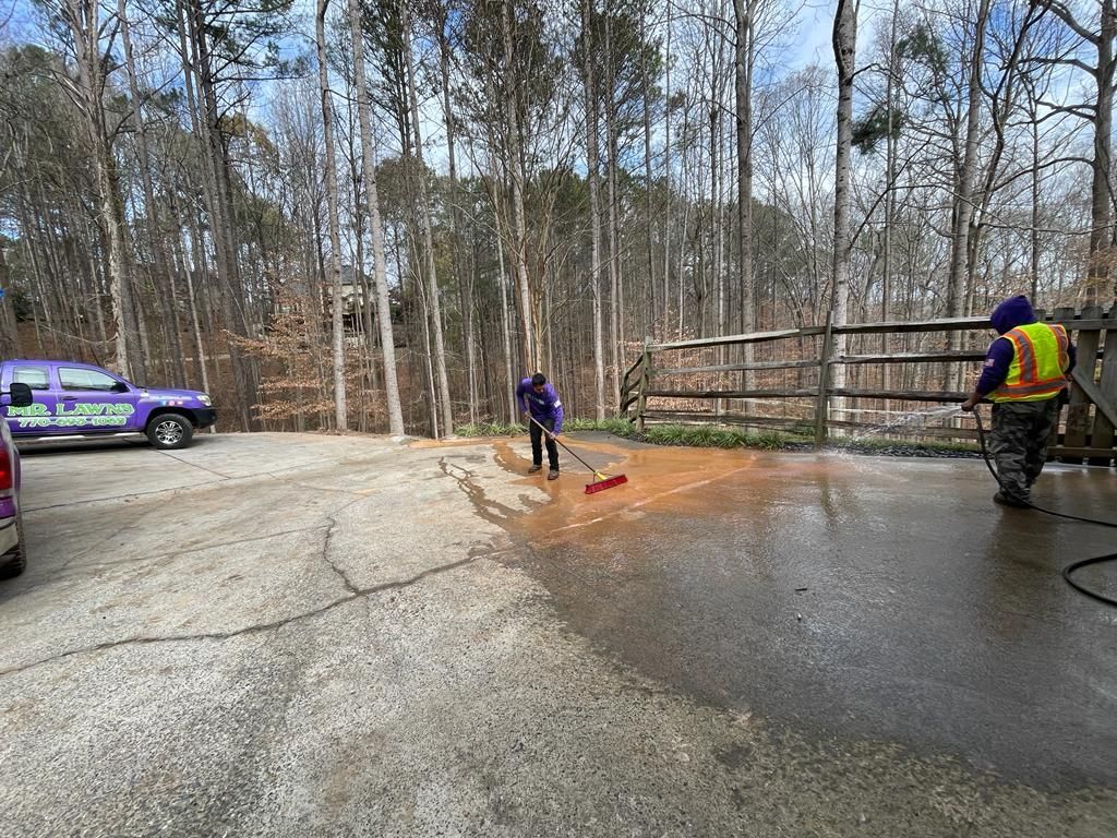 Two people cleaning a driveway. One uses a broom, the other a pressure washer. A purple van sits nearby.