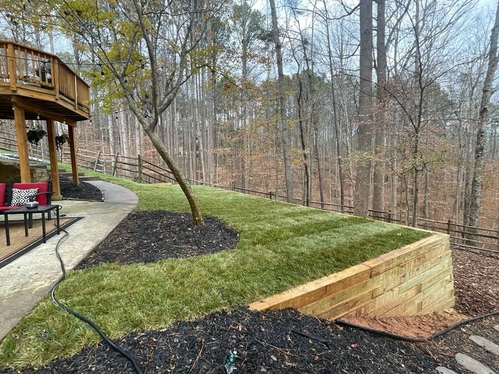 Wooden retaining wall with new sod and mulch near a deck, in a wooded yard.