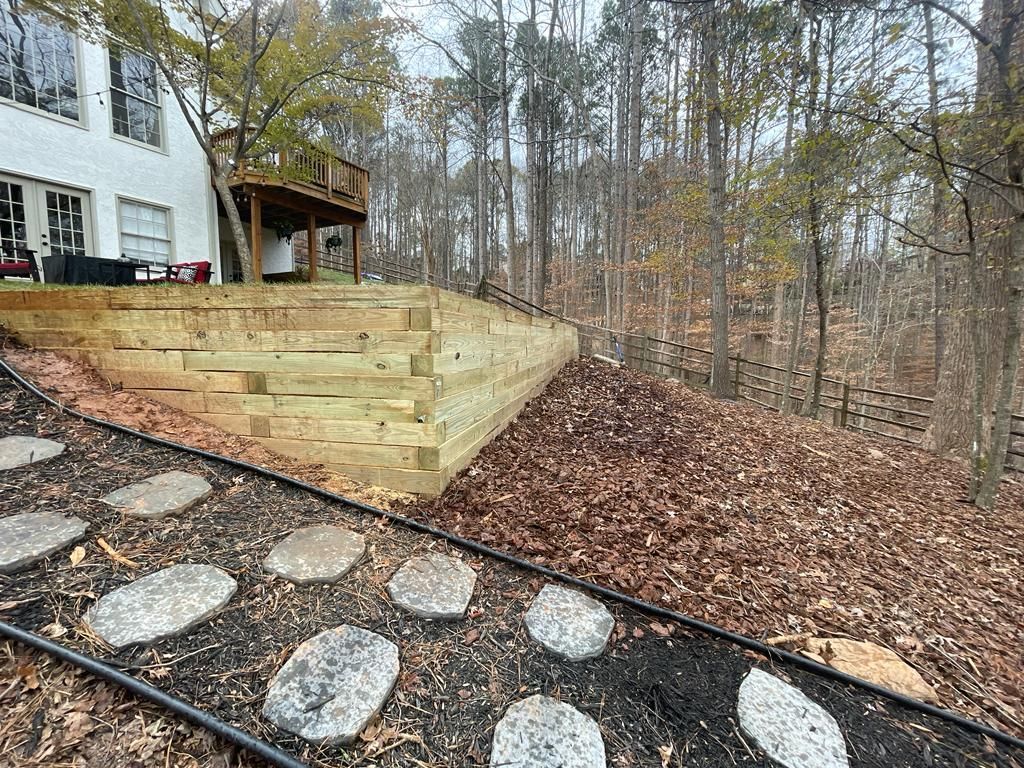 Stone path leads towards a wooden retaining wall and a house with a deck in a wooded area.
