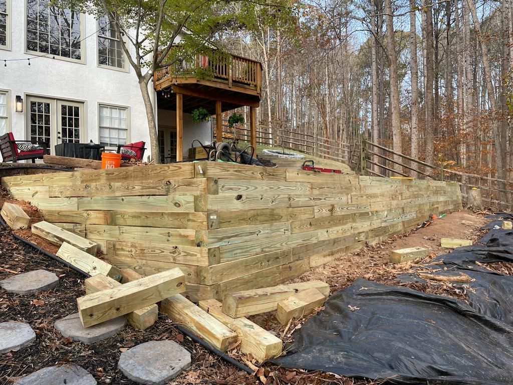 Wooden retaining wall under construction next to a house with a deck in a wooded area.