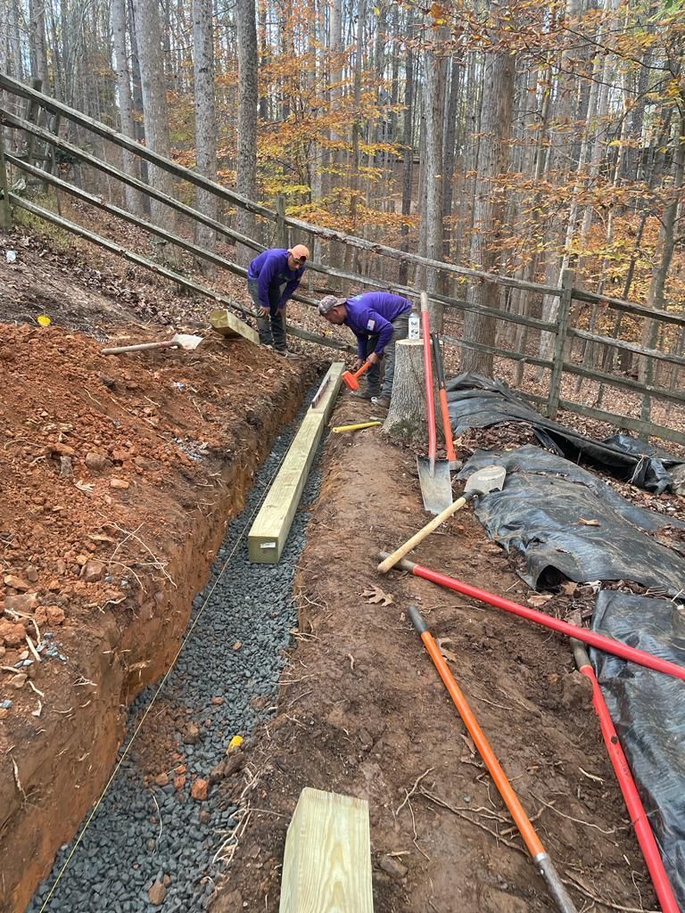 Two workers in purple shirts building a pathway, using shovels and lumber, next to a wooded fence.