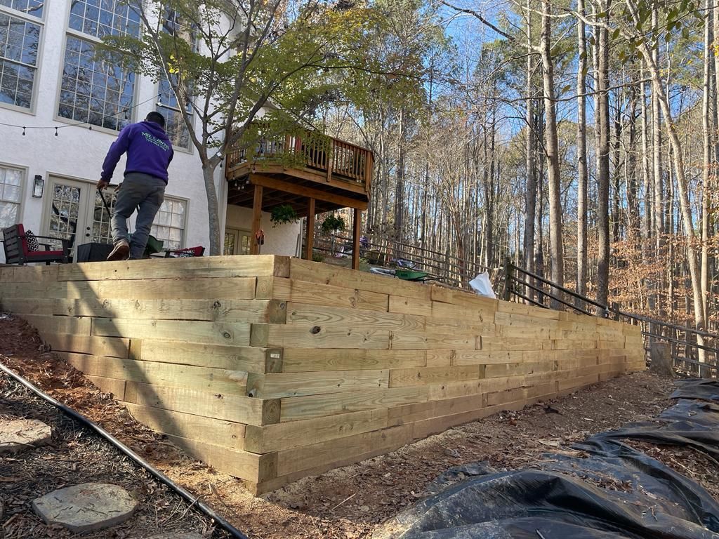 Wooden retaining wall being built near a house. Man in purple shirt works on top of the wall.