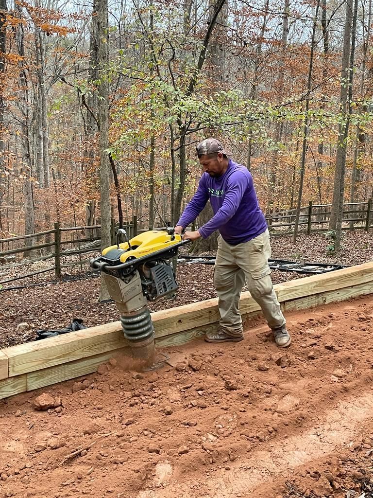 Man operating a soil compactor to flatten a dirt path, near a wooden retaining wall and trees.