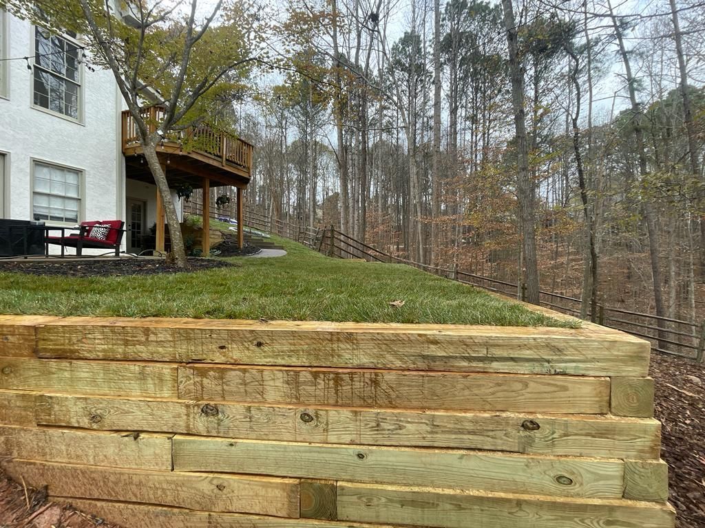 Wooden retaining wall with a grassy hill, leading to a house with a deck and a forested backdrop.
