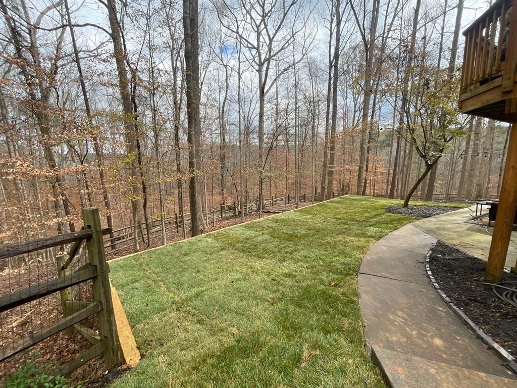 A grassy backyard bordered by a fence and a curved walkway, with a wooded hillside backdrop.