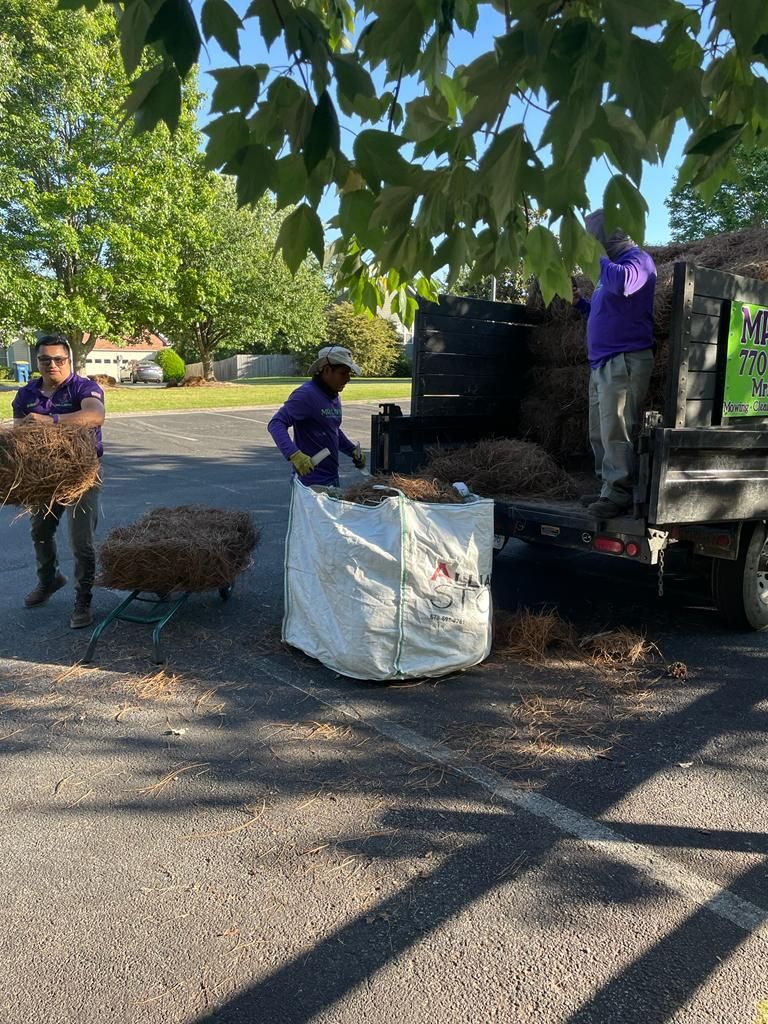 Three people loading a truck with brown debris, one using a cart and a large white bag, outdoors in a parking lot.