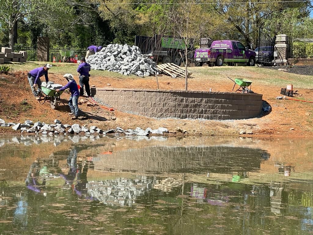 Landscapers building a retaining wall near water. Several workers wear purple shirts and hard hats.