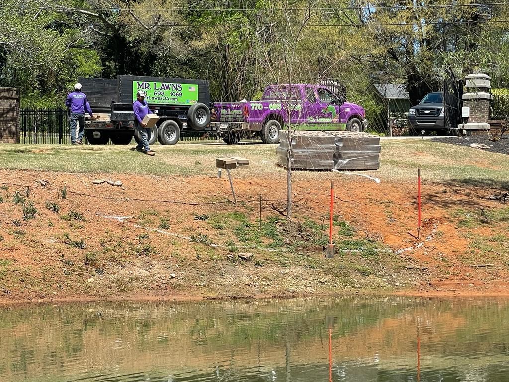 Workers unload debris near a pond. A truck and trailer are visible along with a vehicle covered in purple paint.