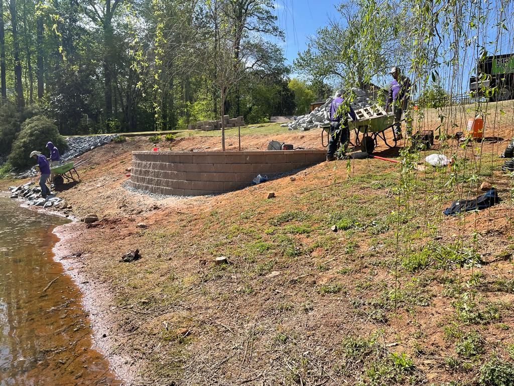 Workers constructing a circular retaining wall on a lake shore. Brown earth, blue sky, and green trees.