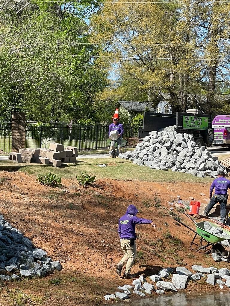 Landscapers in purple shirts and hats work on a hillside, moving rocks and blocks in a sunny outdoor setting.