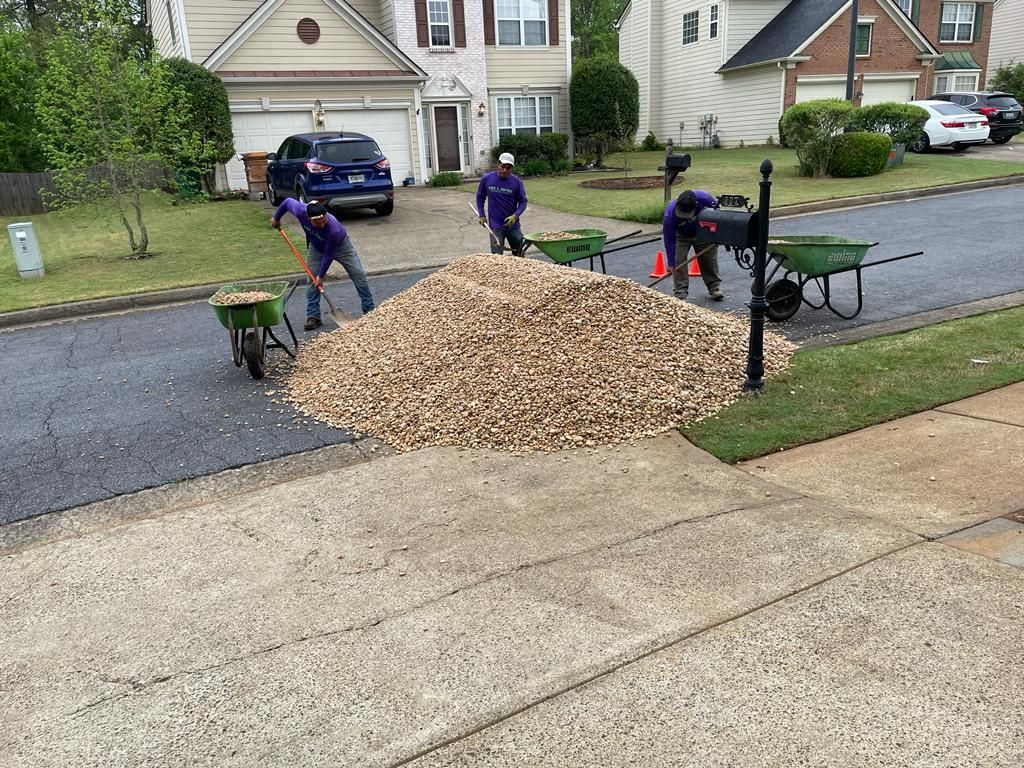 Men shoveling gravel from a large pile on a street into wheelbarrows. Houses and cars in the background.