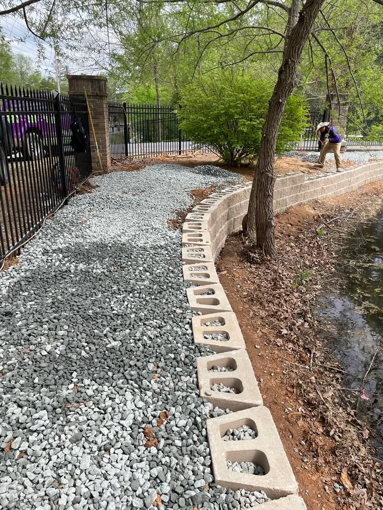 A gravel path with a low concrete retaining wall borders a body of water and a black metal fence; a person works nearby.
