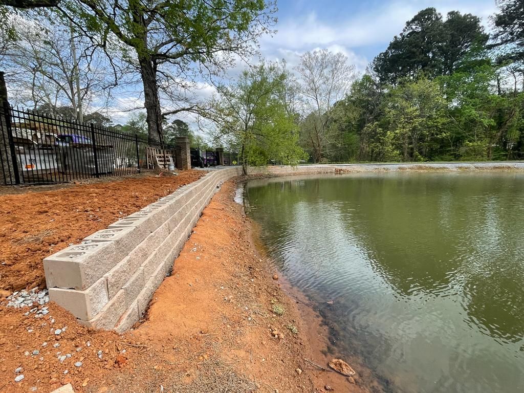 A retaining wall made of concrete blocks borders a pond, with trees in the background.