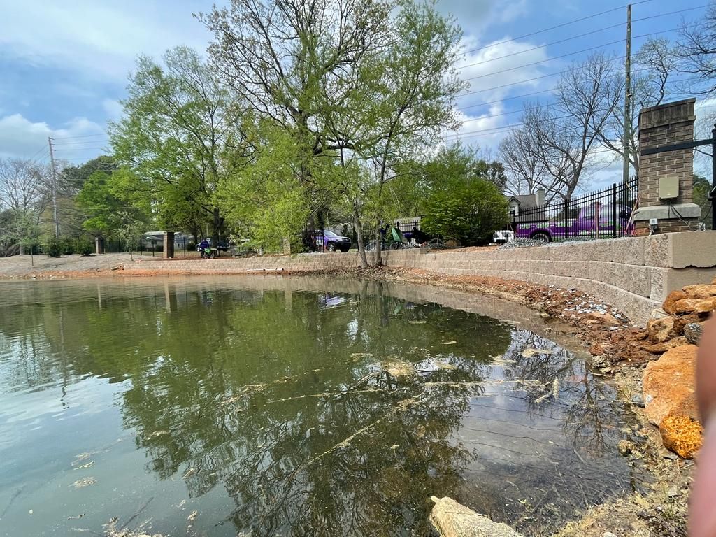 Pond with water reflecting trees and sky; concrete edge, construction debris, and fence in the background.