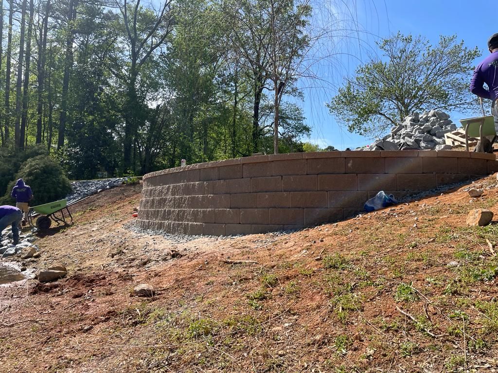 Workers constructing a retaining wall of brown blocks on a hillside. Trees and blue sky are in the background.