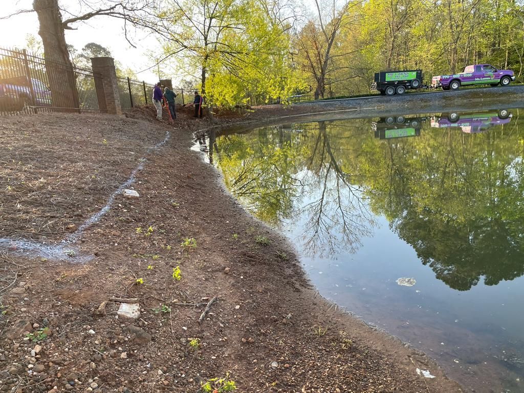 Pond with brown bank, reflections of green trees and vehicles. People are on the bank.