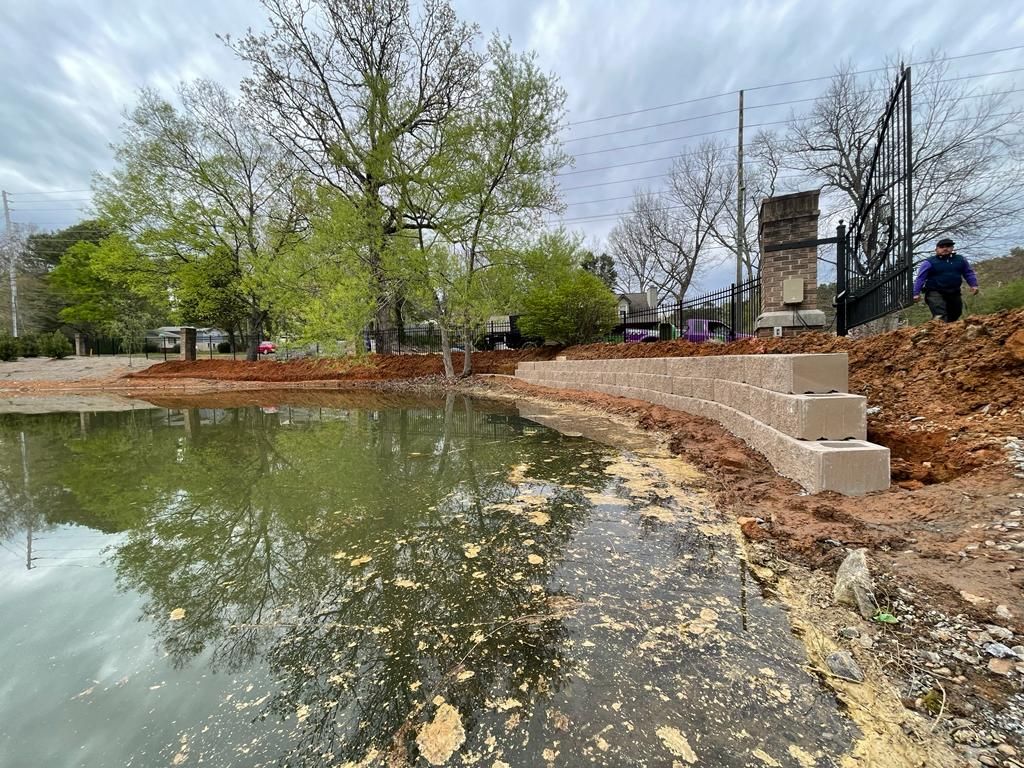 Construction site with a pond and retaining wall. A person walks on a hill near a gate.
