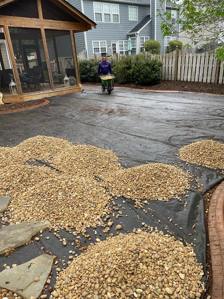 Person using a wheelbarrow to spread gravel on landscaping fabric; backyard setting.