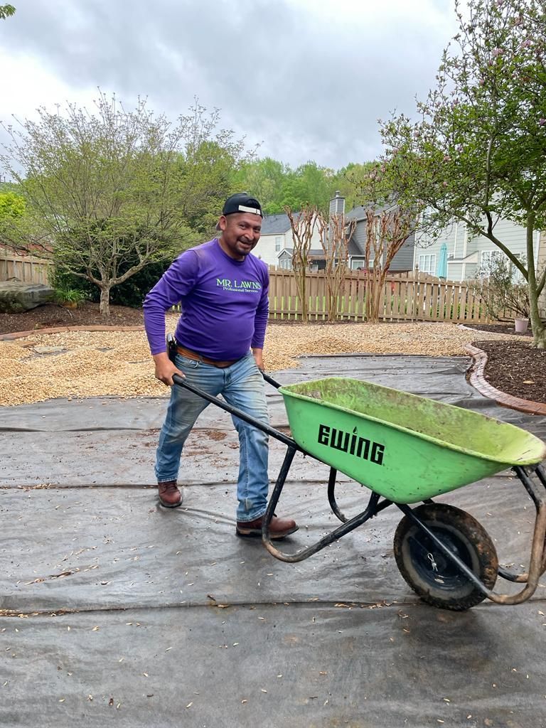 Man in purple shirt pulling a green wheelbarrow filled with grass. Outdoors on a paved surface.