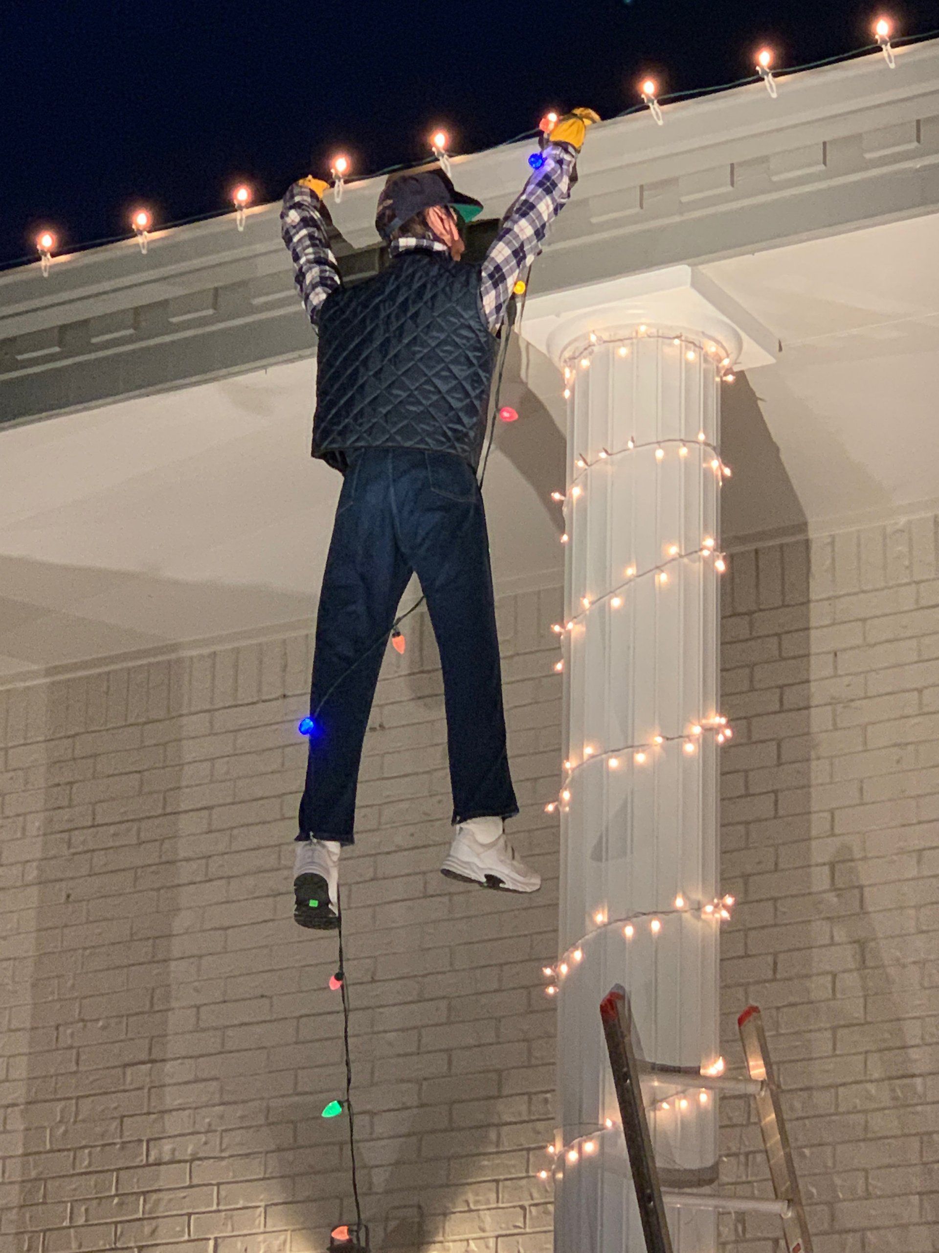 A man is hanging christmas lights from the roof of a building.