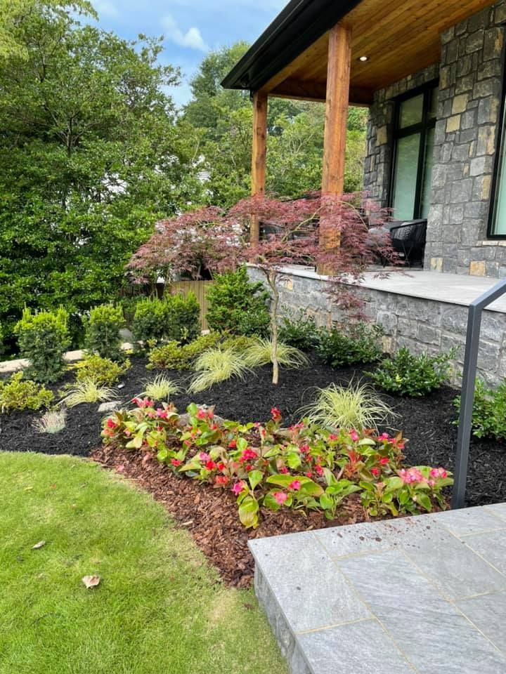 Landscaped yard with colorful flowers, mulch, and a red Japanese maple near a stone house.