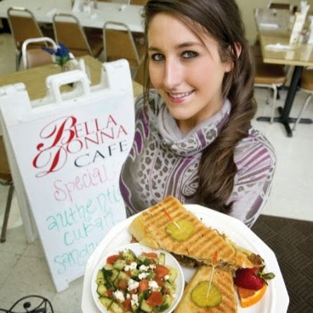 Woman in a café holding a tray with a sandwich, salad, and dessert beside a Bella Donna Cafe sign.