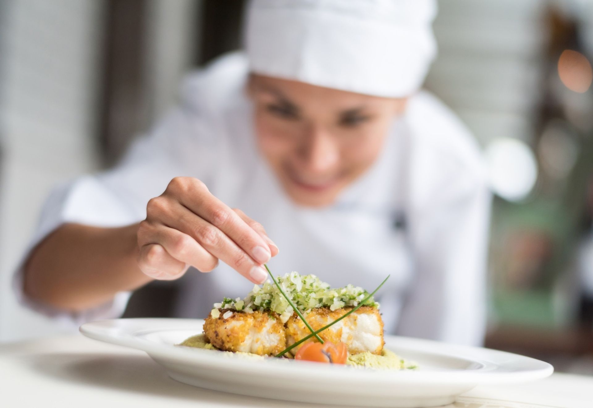 Chef plating a gourmet dish with tweezers on a white plate in a bright kitchen