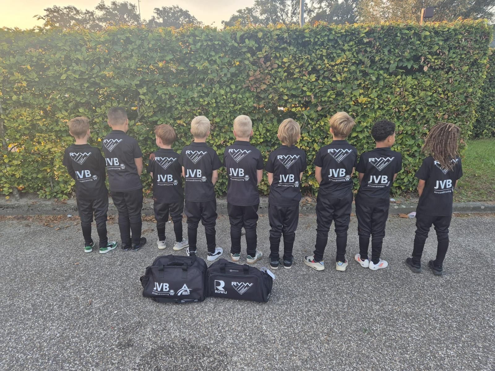 Een groep jonge atleten in zwarte uniformen staat voor een heg, met twee tassen voor zich.