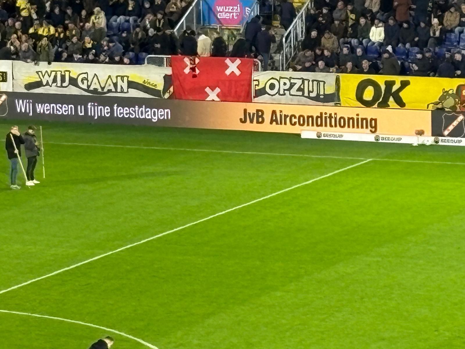 Voetbalveld met toeschouwers, een rode vlag met witte kruizen en reclameborden.