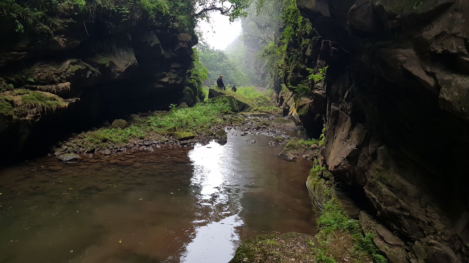Cañón estrecho y rocoso con agua y vegetación, dos figuras caminan hacia la luz.