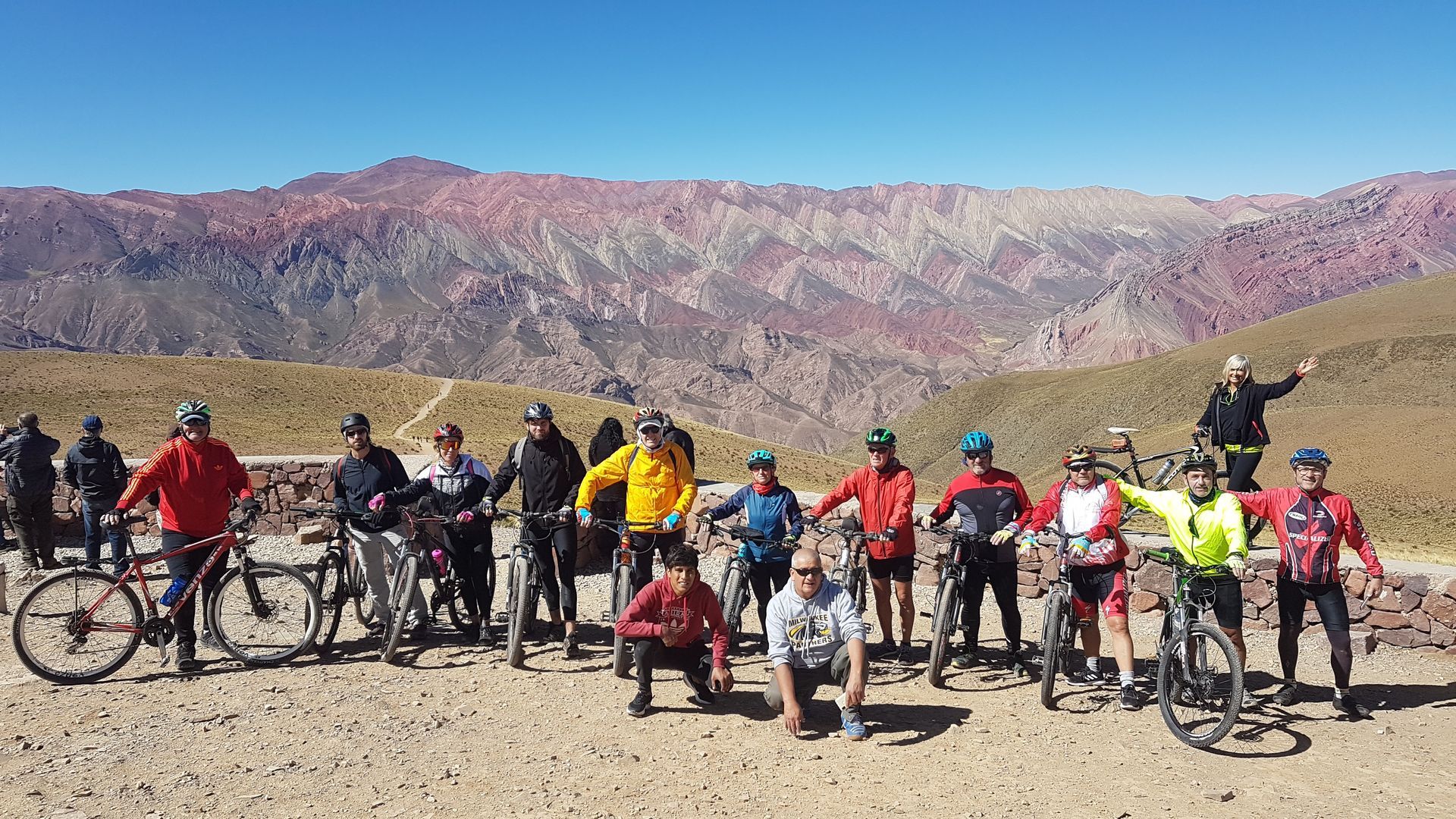 Grupo de ciclistas posando con bicicletas frente a una colorida cadena montañosa bajo un cielo azul.